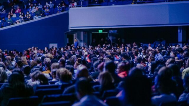 A crowded conference hall with diverse audience attentively listening to a speaker.