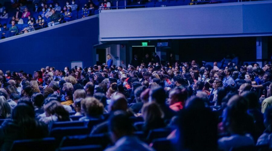 A crowded conference hall with diverse audience attentively listening to a speaker.